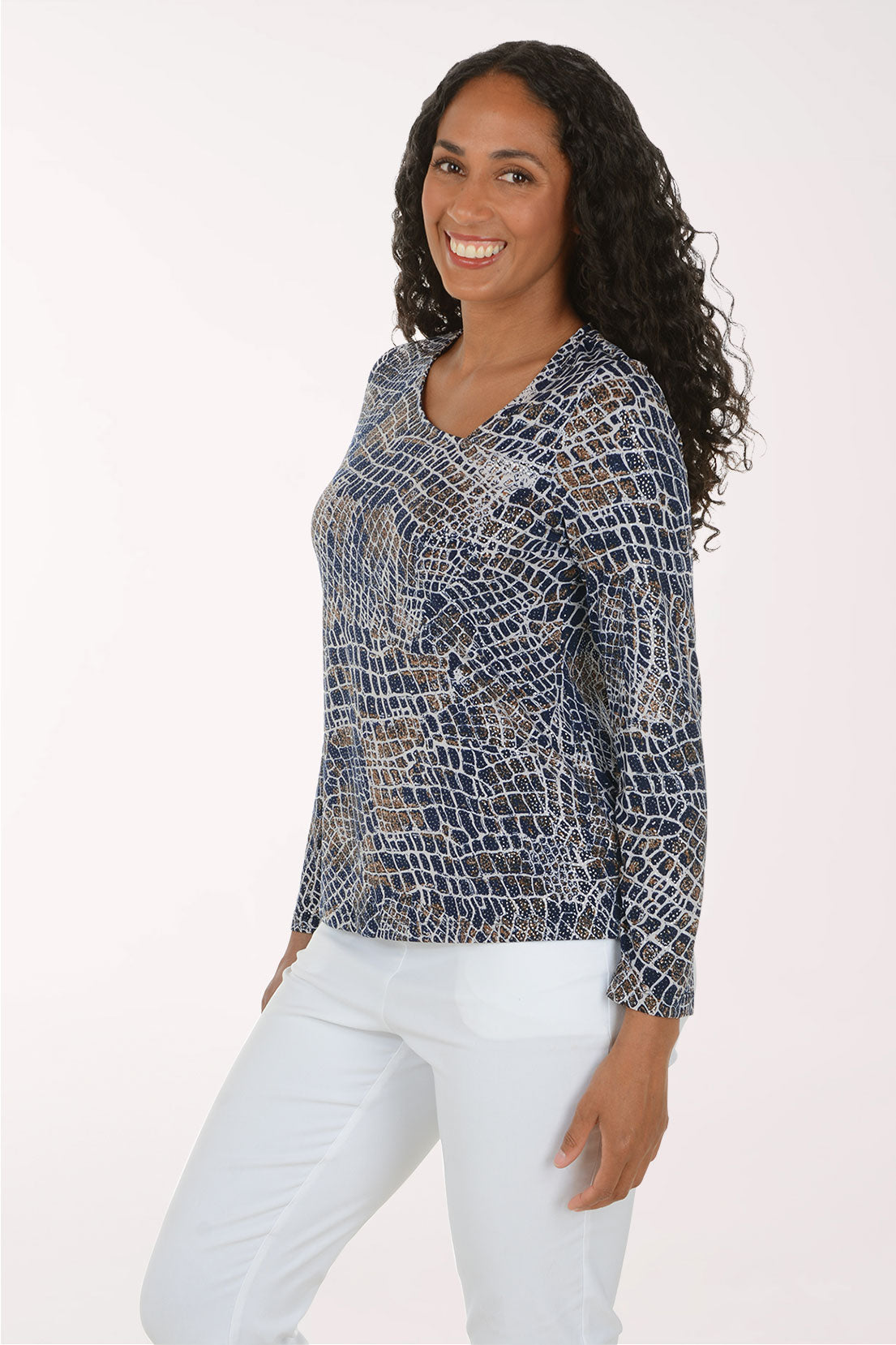 A woman modeling a long-sleeve V-neck top with a blue, white, and beige reptile or snakeskin pattern, paired with white pants, against a plain white studio background.