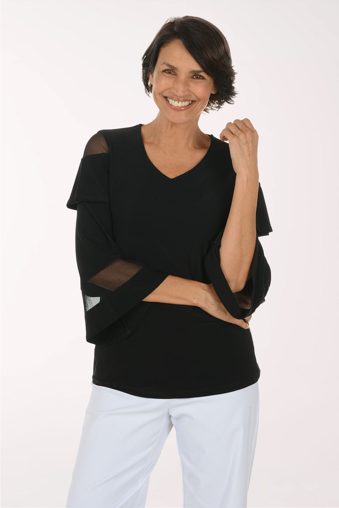 A smiling woman wearing a black V-neck top with sheer mesh shoulder and elbow panels, paired with white pants, photographed against a plain light background.