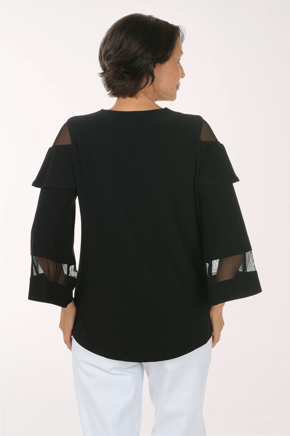 A smiling woman wearing a black V-neck top with sheer mesh shoulder and elbow panels, paired with white pants, photographed against a plain light background.