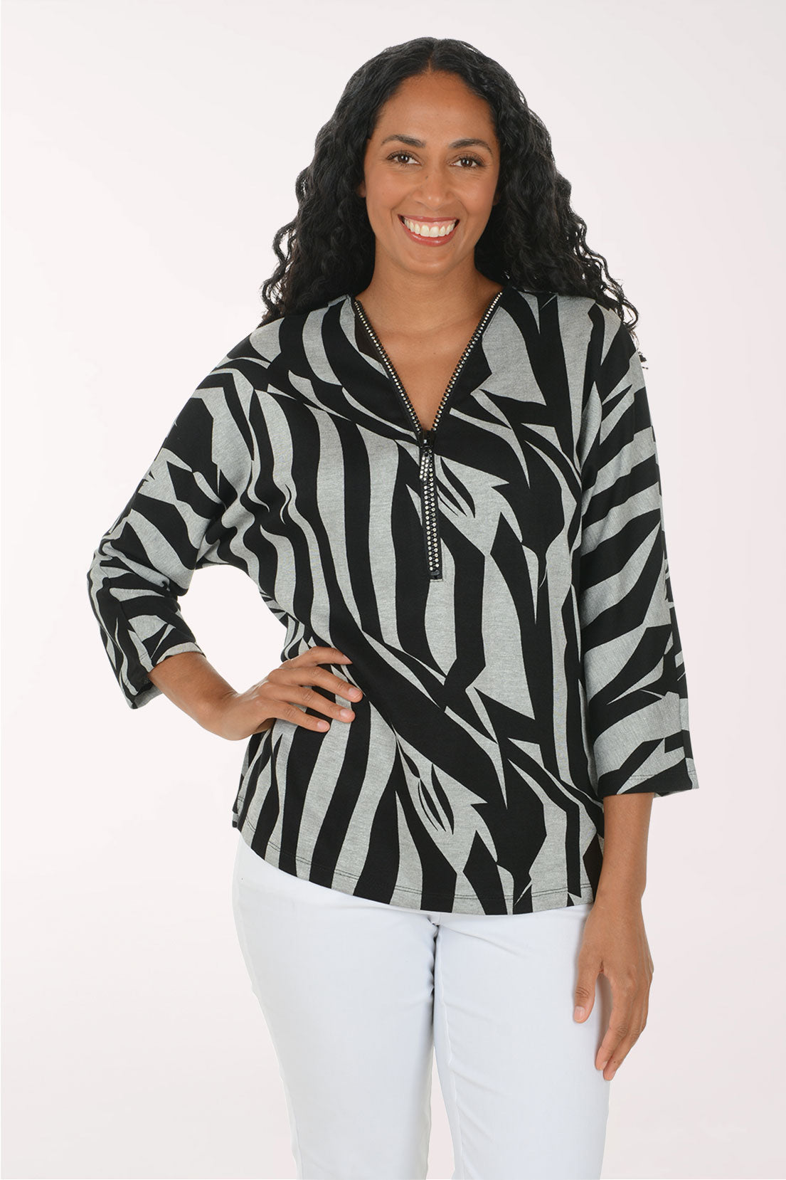 A woman modeling a black and gray abstract patterned top with a half-zip neckline, paired with white pants, against a plain light background.