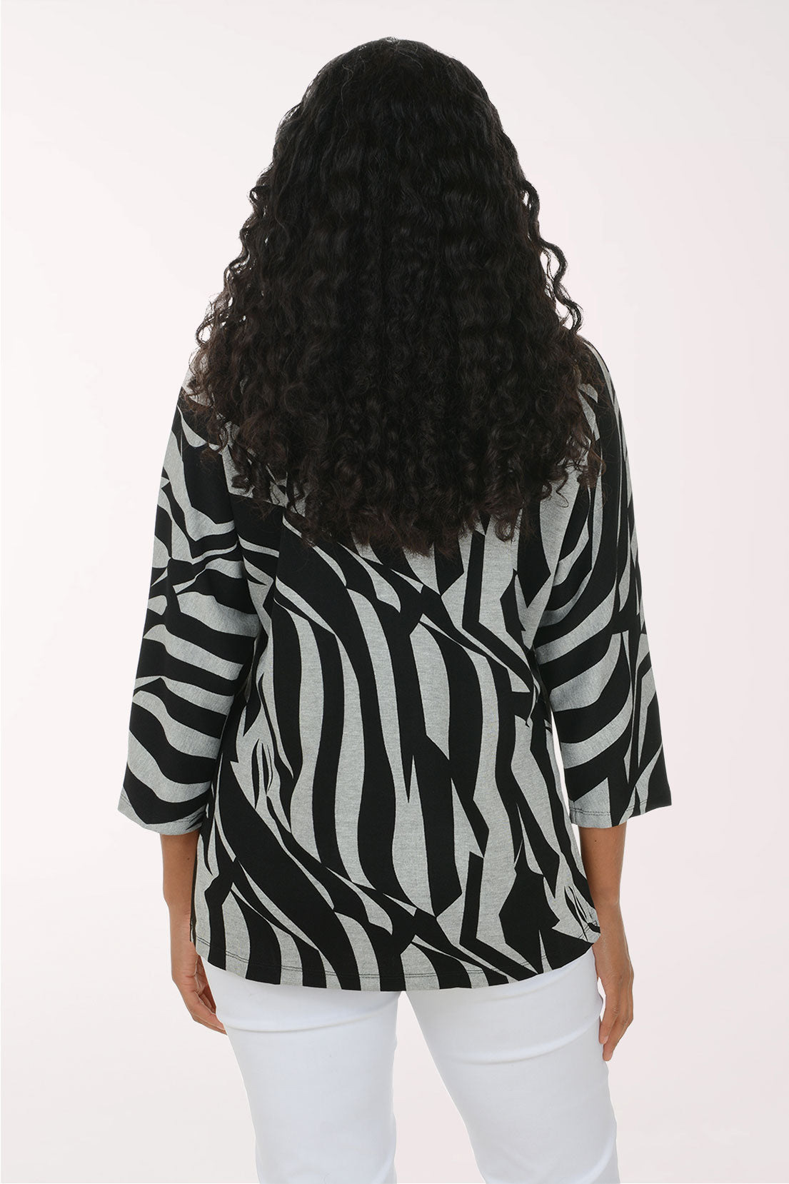 A woman modeling a black and gray abstract patterned top with a half-zip neckline, paired with white pants, against a plain light background.