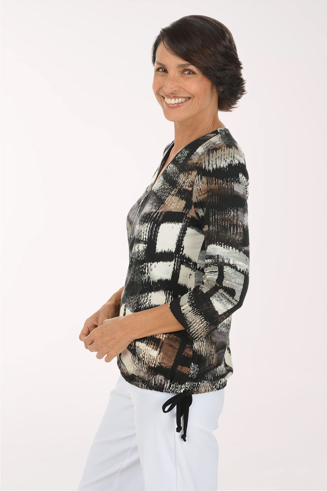 A smiling woman wearing a patterned three-quarter sleeve top with a V-neckline and white pants, standing against a plain light background.