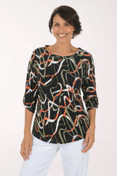 A smiling woman wearing a black three-quarter sleeve top with an abstract wavy pattern in white, orange, and olive green, paired with white pants, against a plain white studio background.