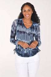 A woman modeling a long-sleeve V-neck top with a blue and gray tie-dye pattern, paired with white pants, against a plain white background.