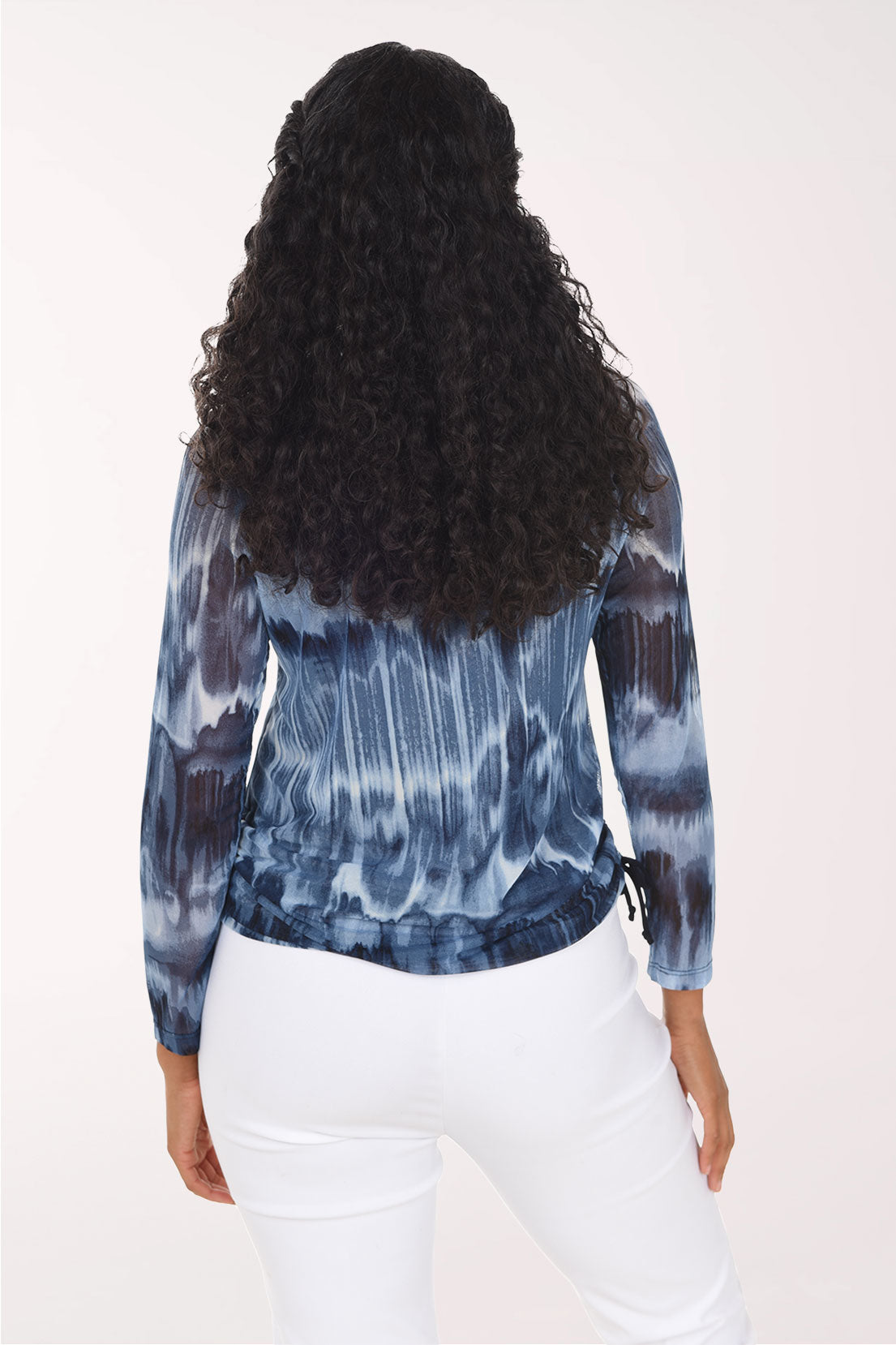 A woman modeling a long-sleeve V-neck top with a blue and gray tie-dye pattern, paired with white pants, against a plain white background.