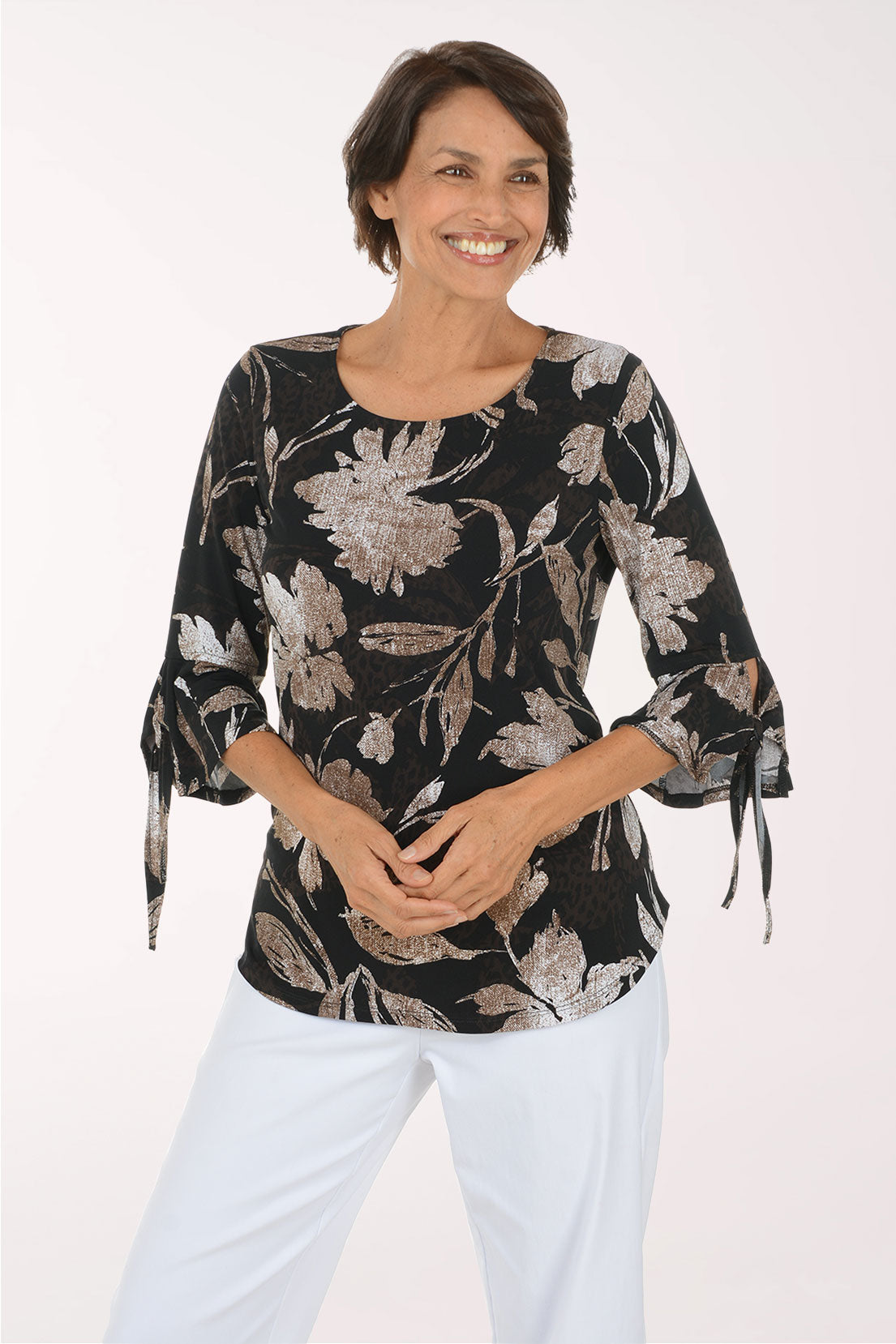 A woman modeling a black three-quarter sleeve blouse with a beige and gray floral pattern, paired with white pants, against a plain white studio background.