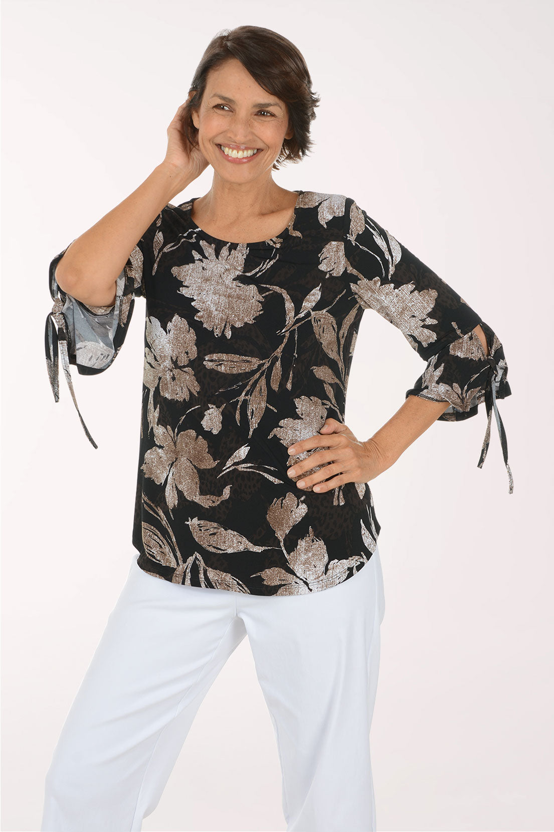 A woman modeling a black three-quarter sleeve blouse with a beige and gray floral pattern, paired with white pants, against a plain white studio background.
