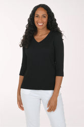 A woman with long, curly black hair wearing a black ribbed three-quarter sleeve V-neck top and white pants, standing against a plain light background.