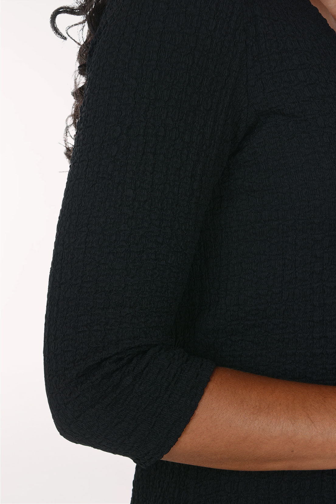 Close-up of a person wearing a black textured three-quarter sleeve top, showing the ribbed fabric and part of the arm against a plain light background.