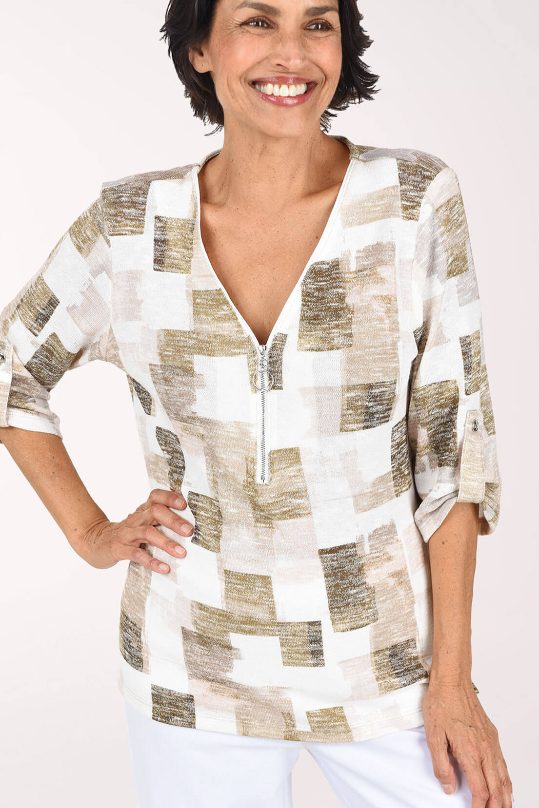 Close up of model wearing a white, beige, and blush abstract print knit top with a zip V-neck and roll-tab sleeves, paired with white pants.