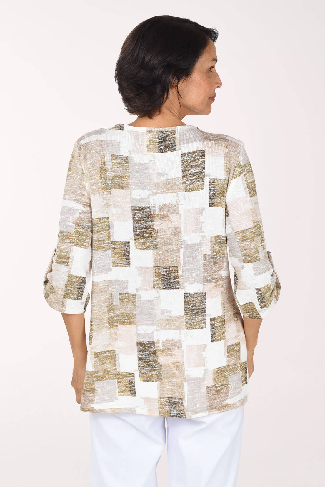 Back shot of model wearing a white, beige, and blush abstract print knit top with a zip V-neck and roll-tab sleeves, paired with white pants.