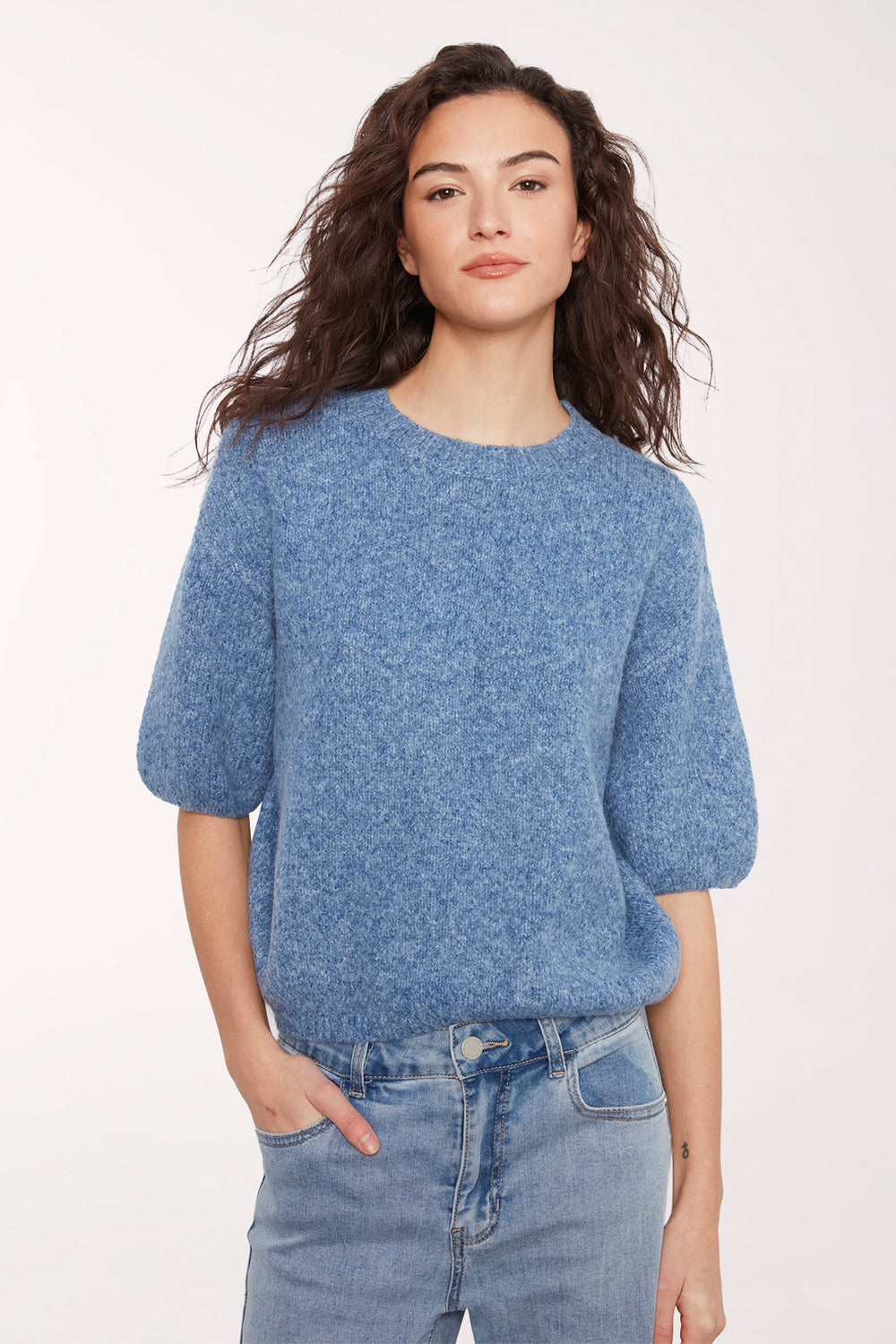 A woman wearing a textured, heathered blue short-sleeve knit top and light blue jeans against a plain white studio background.