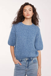 A woman wearing a textured, heathered blue short-sleeve knit top and light blue jeans against a plain white studio background.