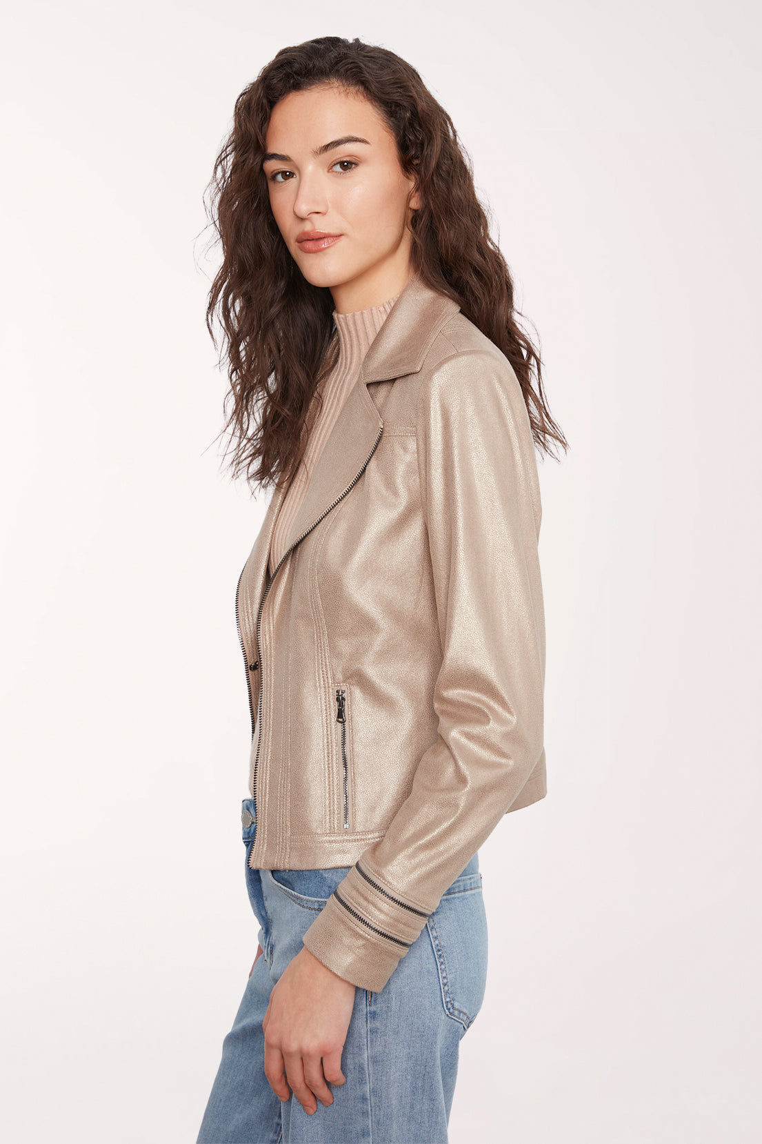A woman wearing a metallic champagne beige faux leather moto jacket over a ribbed turtleneck and blue jeans, photographed against a clean white studio background.