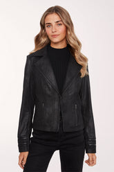 A woman modeling a black faux-leather moto jacket with silver zippers, worn over a black ribbed top and black pants, against a plain white studio background.