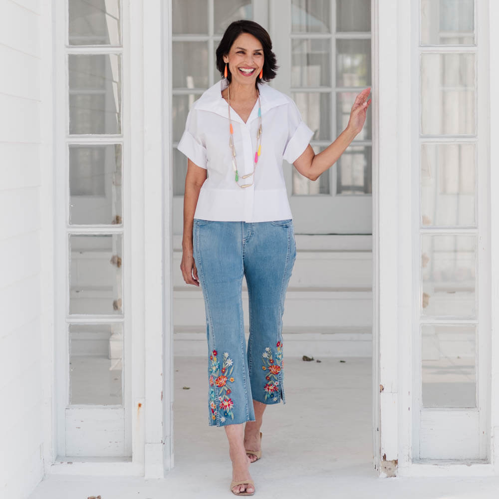 Woman wearing a white blouse and blue jeans with floral embroidery, standing in a bright room.