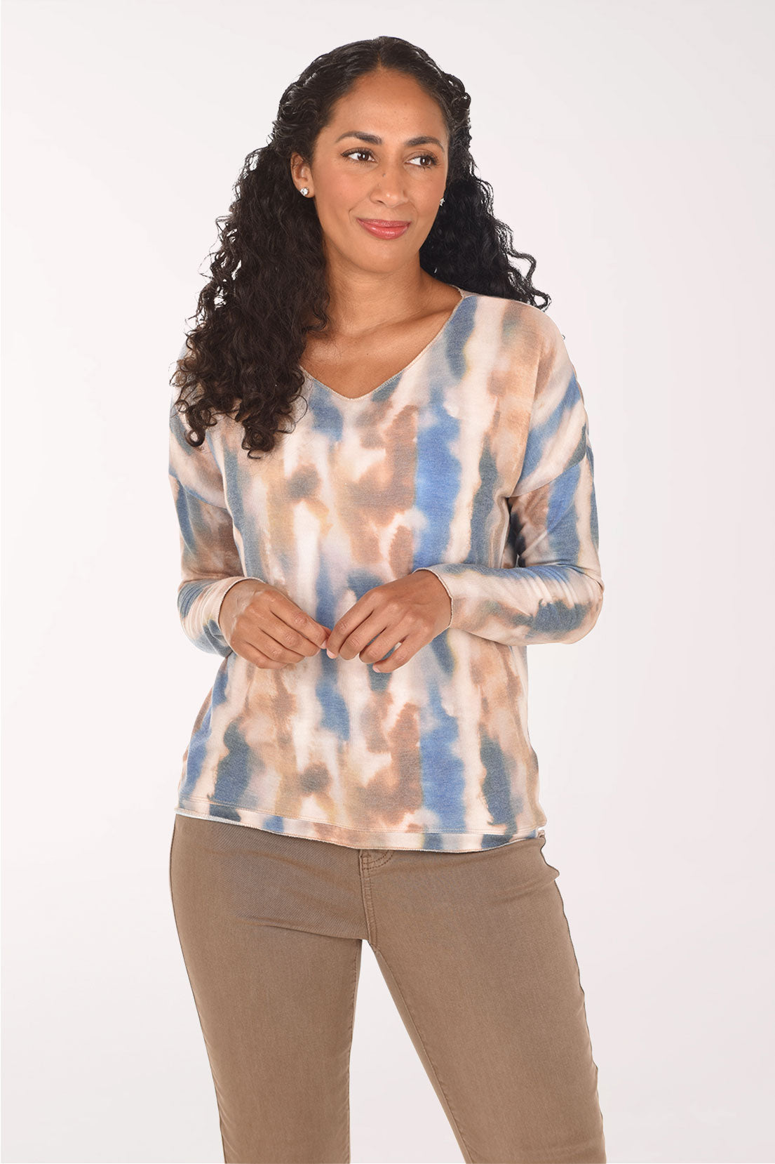 A woman with long curly black hair wearing a long-sleeve V-neck top with a blue, beige, and brown tie-dye pattern, paired with brown pants, against a plain white background.