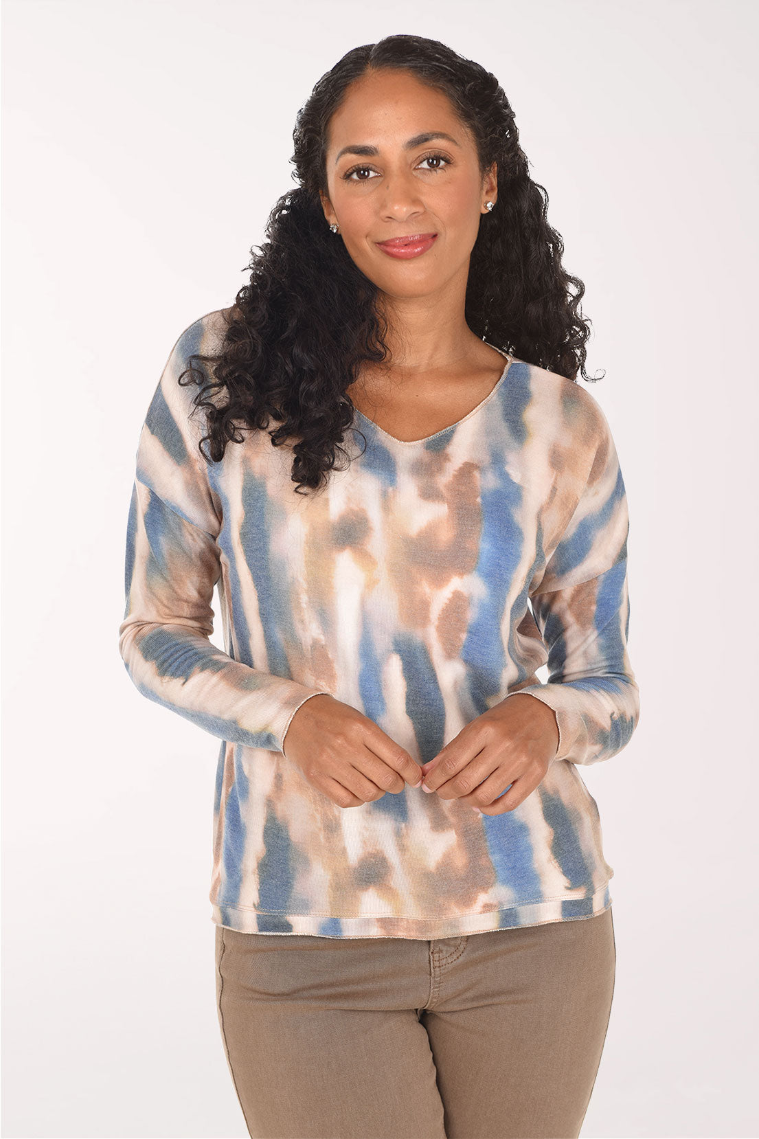 A woman with long curly black hair wearing a long-sleeve V-neck top with a blue, beige, and brown tie-dye pattern, paired with brown pants, against a plain white background.