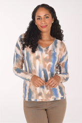 A woman with long curly black hair wearing a long-sleeve V-neck top with a blue, beige, and brown tie-dye pattern, paired with brown pants, against a plain white background.