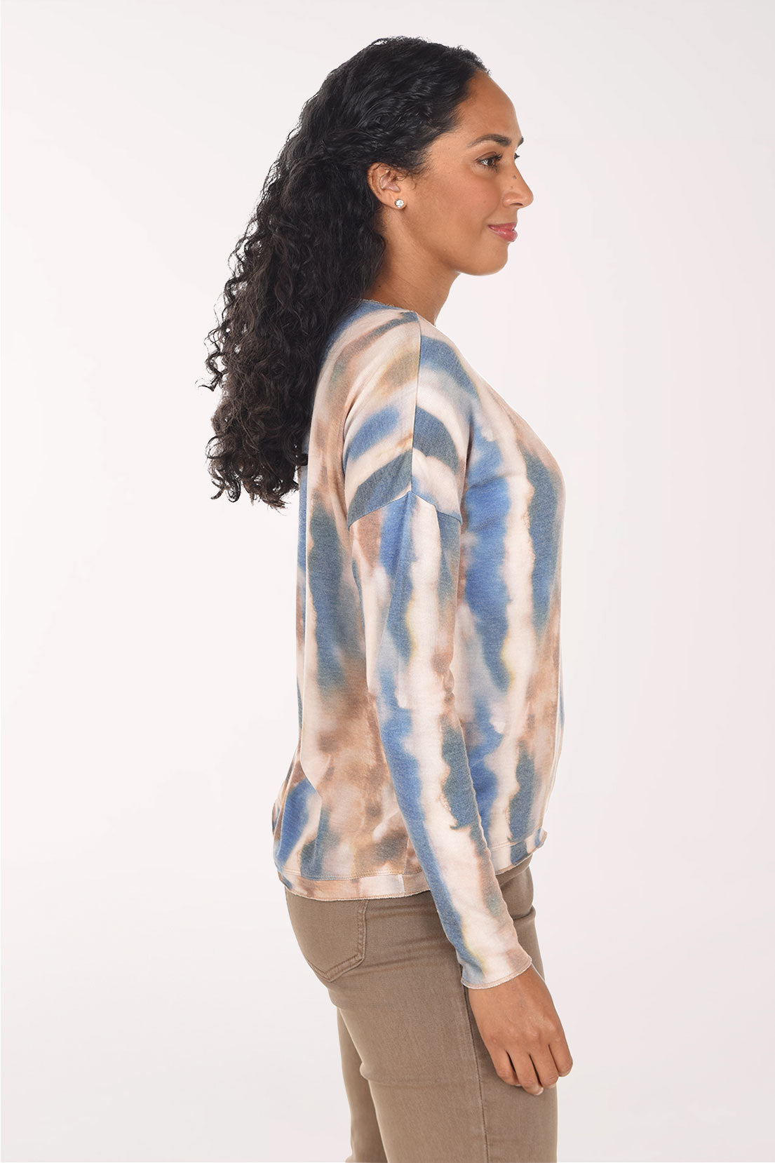 A woman with long curly black hair wearing a long-sleeve V-neck top with a blue, beige, and brown tie-dye pattern, paired with brown pants, against a plain white background.