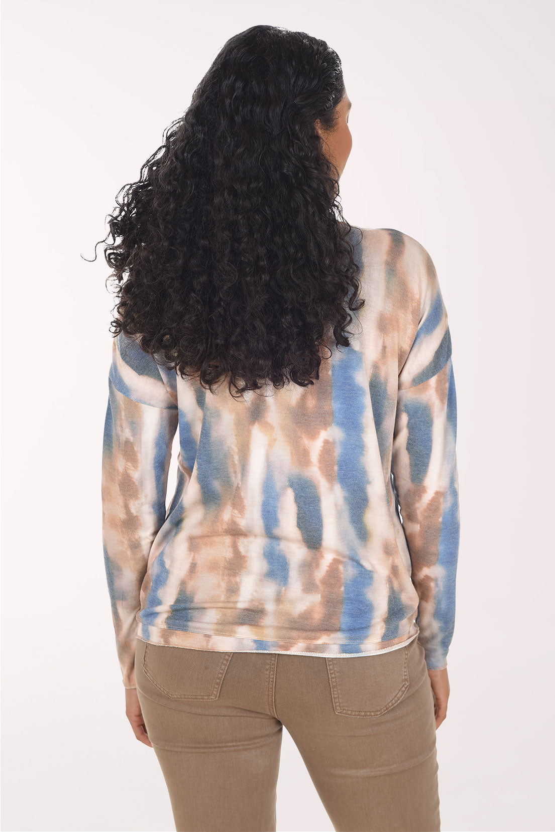 A woman with long curly black hair wearing a long-sleeve V-neck top with a blue, beige, and brown tie-dye pattern, paired with brown pants, against a plain white background.
