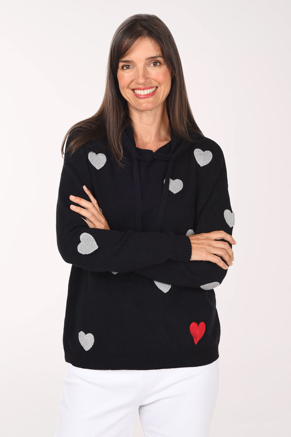 A woman wearing a black hoodie sweater covered in grey boucle hearts with one red heart, arms crossed, with white pants.