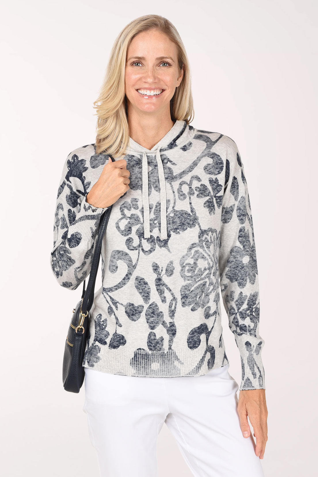 A woman wearing an ivory and grey hoodie with a large navy and charcoal floral print, carrying a black crossbody bag, with white pants.