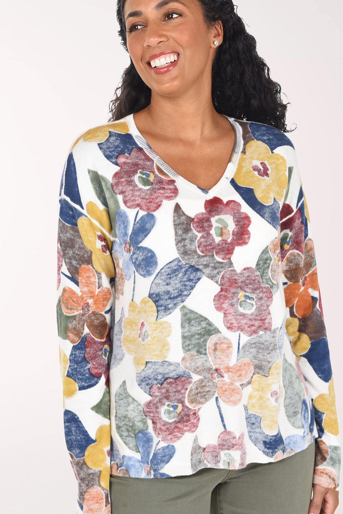 A woman wearing a white long-sleeve V-neck top with a colorful floral print in blue, red, yellow, and orange, paired with olive green pants, against a plain light background.