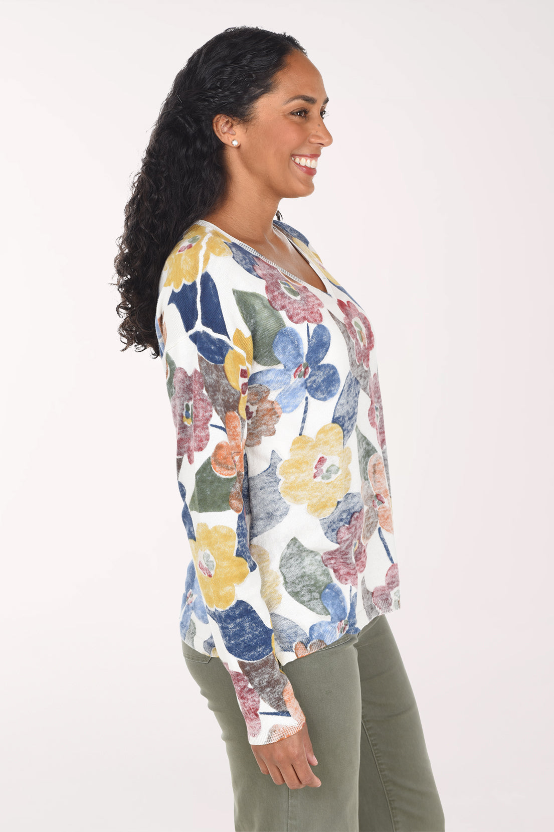 A woman wearing a white long-sleeve V-neck top with a colorful floral print in blue, red, yellow, and orange, paired with olive green pants, against a plain light background.