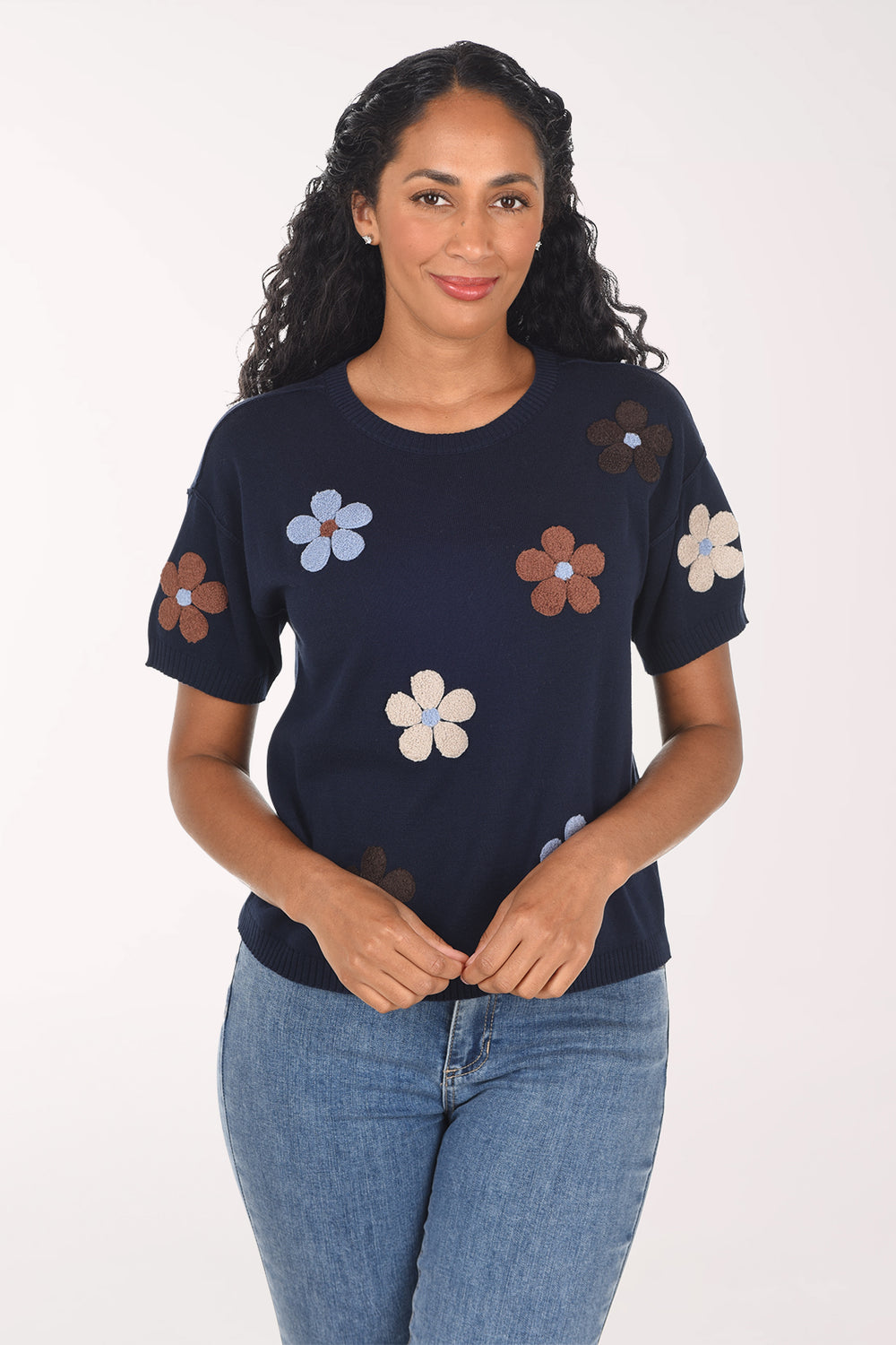 A woman modeling a navy blue short-sleeve top with embroidered flowers in brown, light blue, and cream, paired with blue jeans against a plain white background.