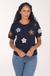A woman modeling a navy blue short-sleeve top with embroidered flowers in brown, light blue, and cream, paired with blue jeans against a plain white background.