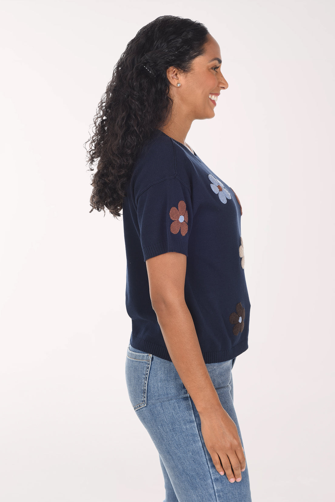 A woman modeling a navy blue short-sleeve top with embroidered flowers in brown, light blue, and cream, paired with blue jeans against a plain white background.