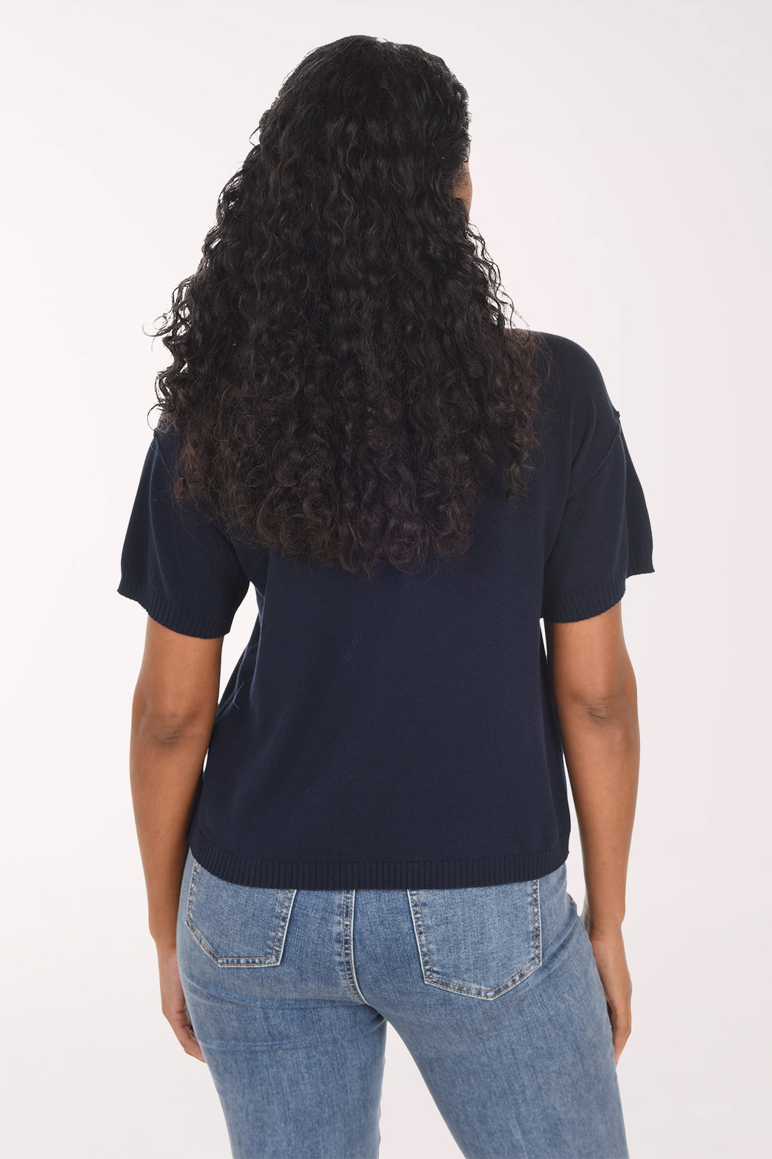 A woman modeling a navy blue short-sleeve top with embroidered flowers in brown, light blue, and cream, paired with blue jeans against a plain white background.