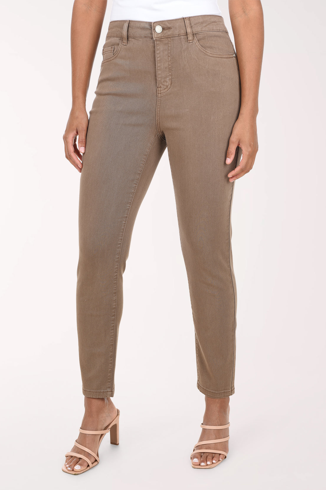 A model wearing a pair of slim-fit, cropped ankle-length jeans in a muted taupe-brown color, paired with beige strappy high-heeled sandals against a plain light background.