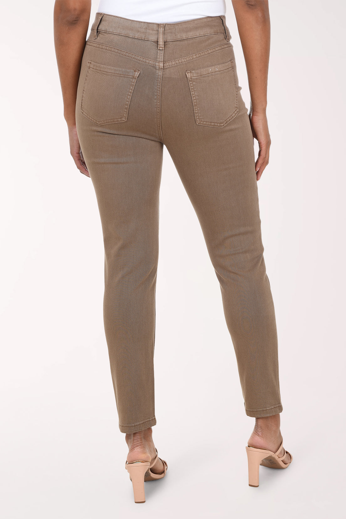 A model wearing a pair of slim-fit, cropped ankle-length jeans in a muted taupe-brown color, paired with beige strappy high-heeled sandals against a plain light background.