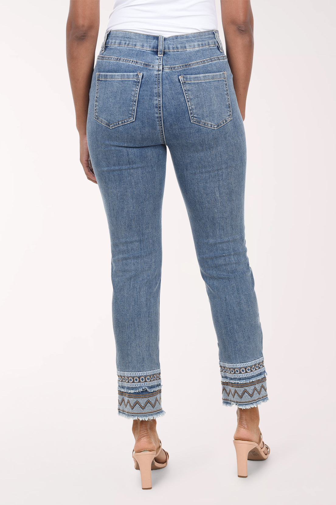 A model wearing a pair of medium-wash blue denim jeans with a geometric pattern trim at the rolled cuffs, paired with a white top and beige strappy heels, against a plain light background.