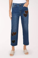 A front view of medium wash straight leg jeans with dark brown and black heart-shaped patches on the legs, worn with nude sandals.