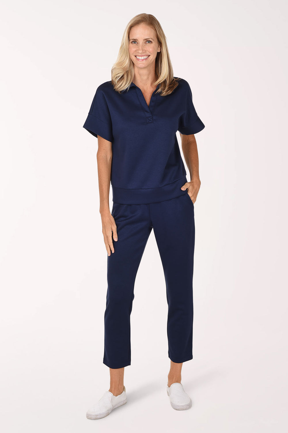 Full-length front view of a woman in navy Jet Blue scuba knit straight leg ankle pants with a smocked waistband, worn with the matching navy scuba polo top and white sneakers.