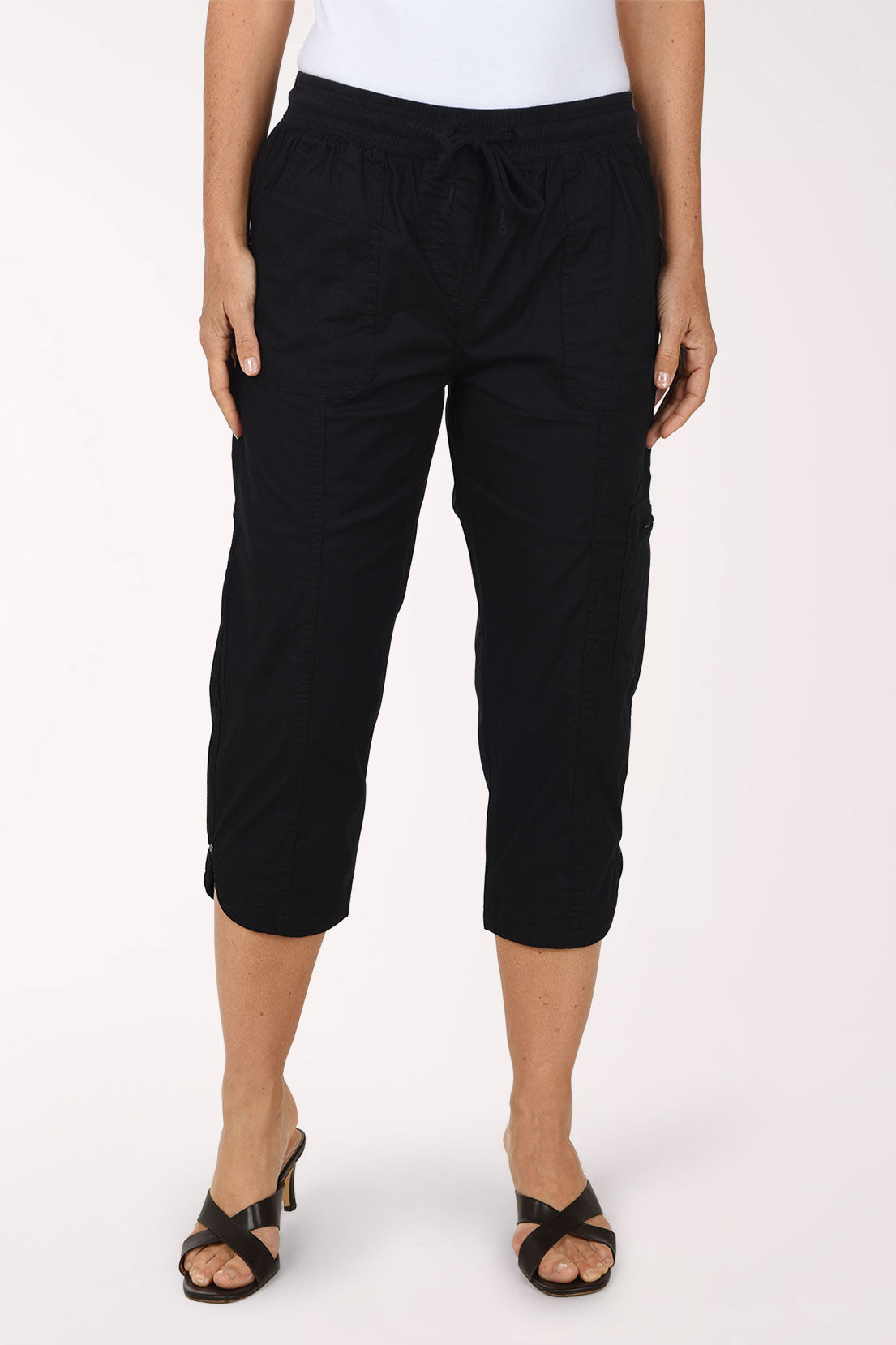 Front view of black cotton poplin capri pants with a rib knit drawstring waistband, front utility pockets, a zip cargo pocket on the left leg and a tulip hem with rivets, worn just below the knee with a white top and black heeled sandals.