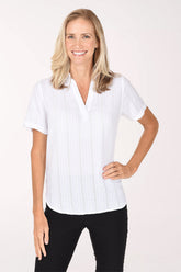 A smiling blonde woman wearing a white short-sleeve button-front blouse with subtle vertical texture and black pants, standing against a plain light background.