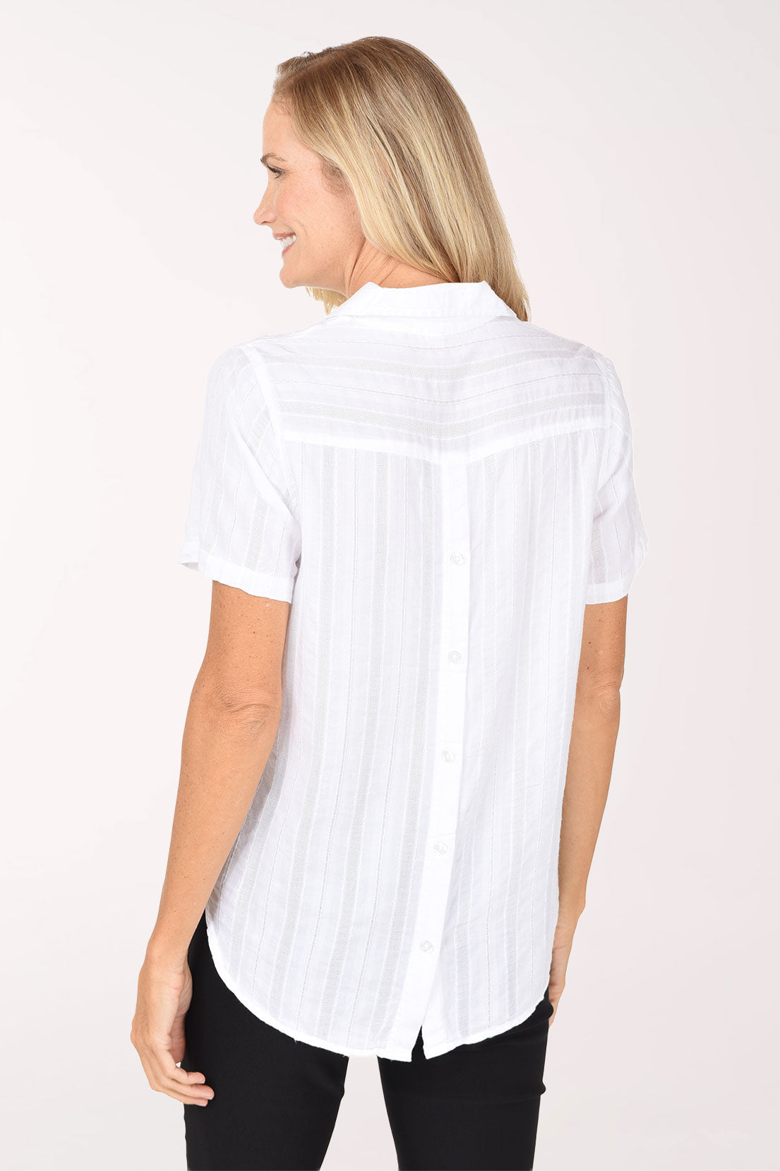 A smiling blonde woman wearing a white short-sleeve button-front blouse with subtle vertical texture and black pants, standing against a plain light background.