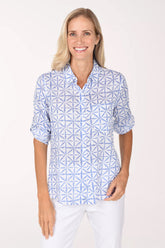 A smiling blonde woman wearing a light blue and white geometric patterned button-down shirt, posing against a solid off-white studio background.