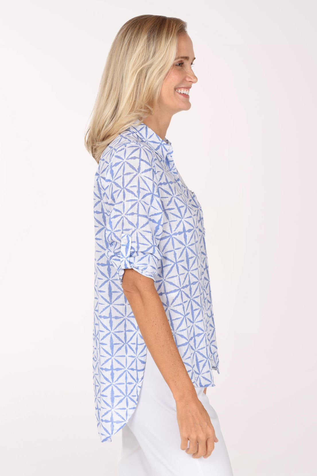 A smiling blonde woman wearing a light blue and white geometric patterned button-down shirt, posing against a solid off-white studio background.