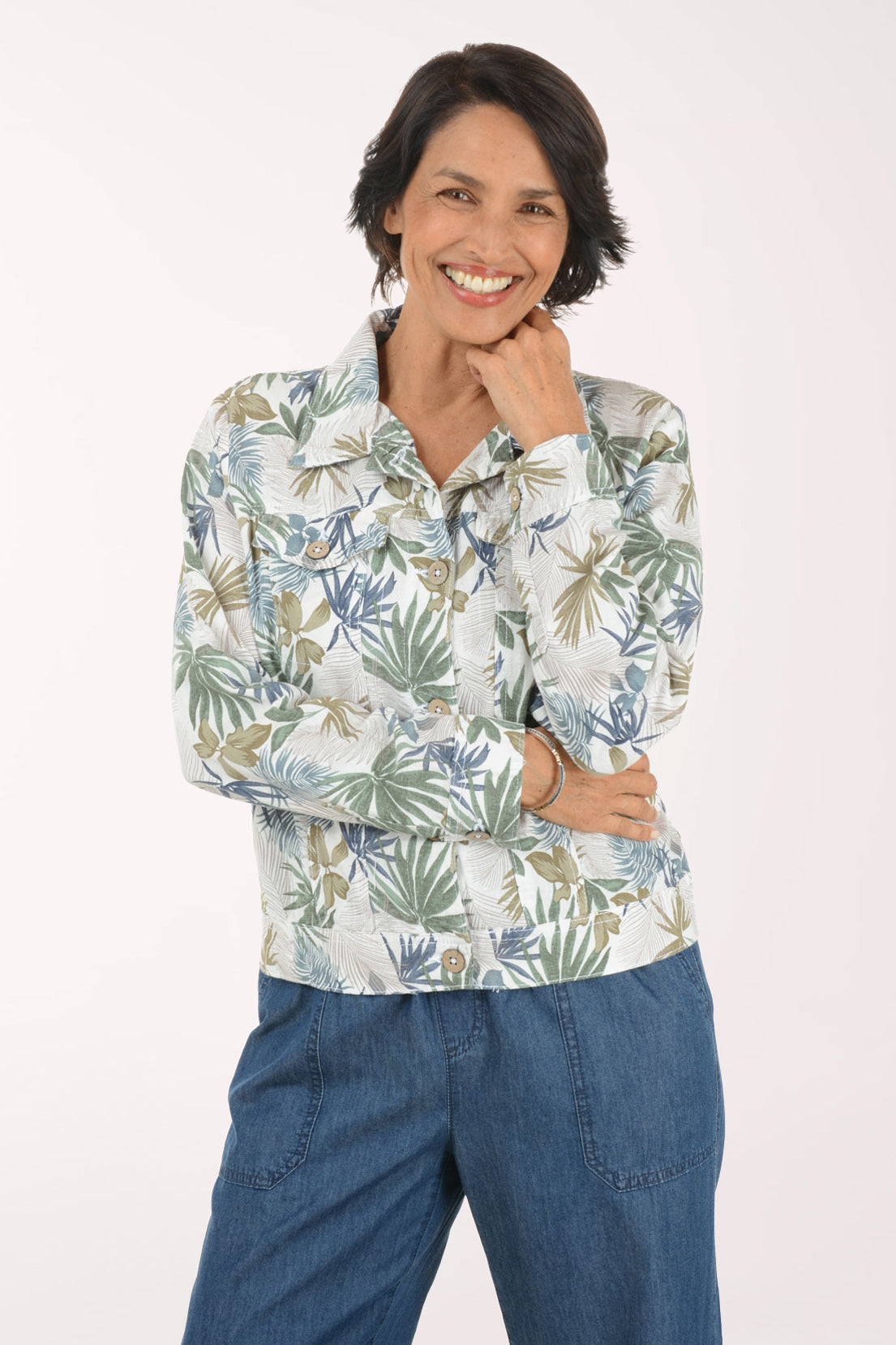 A woman wearing a long-sleeve button-up shirt with a blue, green, and beige palm leaf pattern, paired with blue jeans, posing against a solid white background.