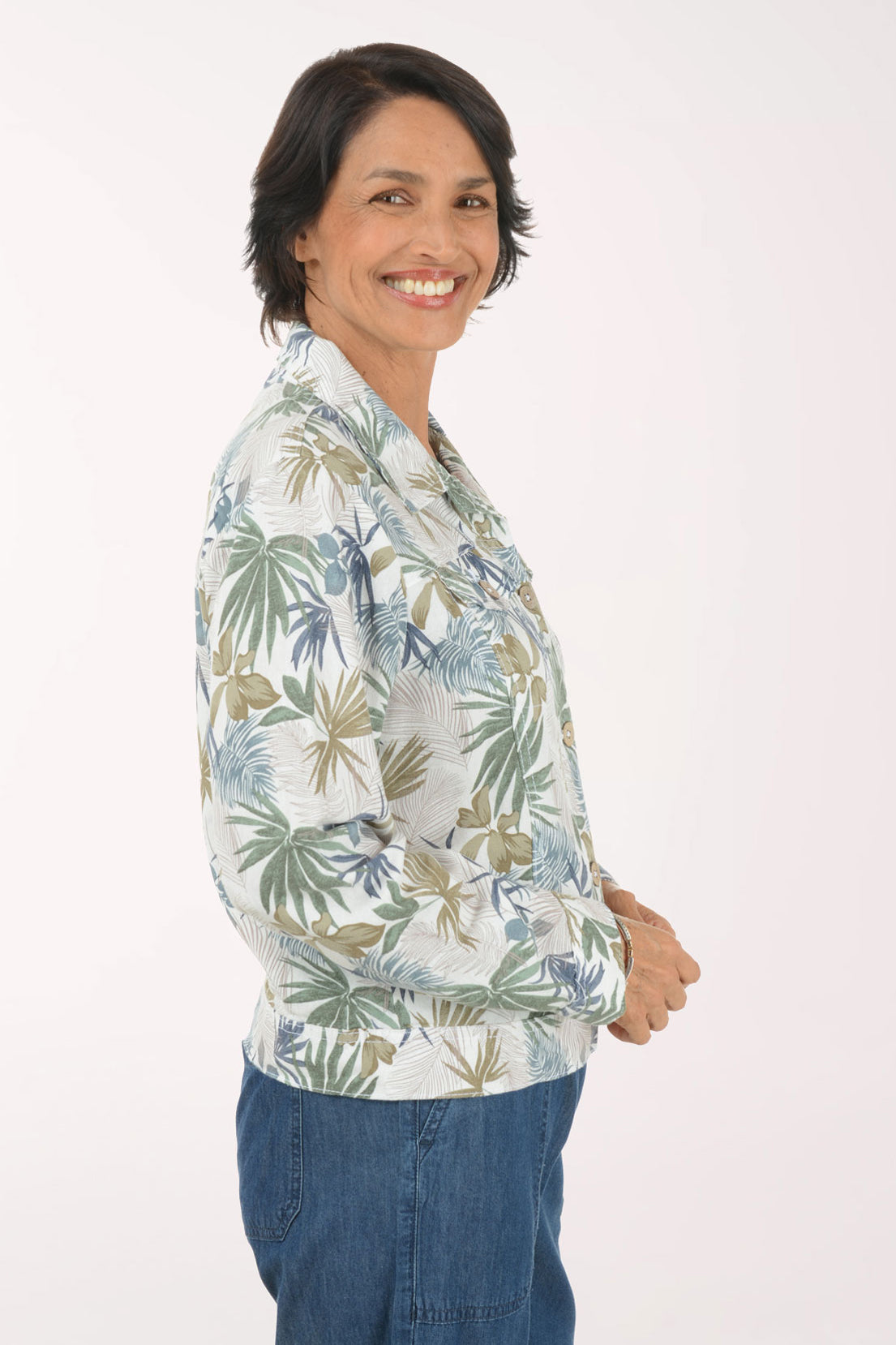 A woman wearing a long-sleeve button-up shirt with a blue, green, and beige palm leaf pattern, paired with blue jeans, posing against a solid white background.