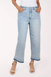 A person wearing light-wash cropped denim jeans with a straight-to-wide leg, shown from the waist down against a plain light background. The model has their hands in the pockets and is wearing beige heeled sandals.