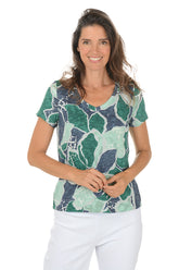 Woman wearing a green and blue patterned top with white pants on a white background.