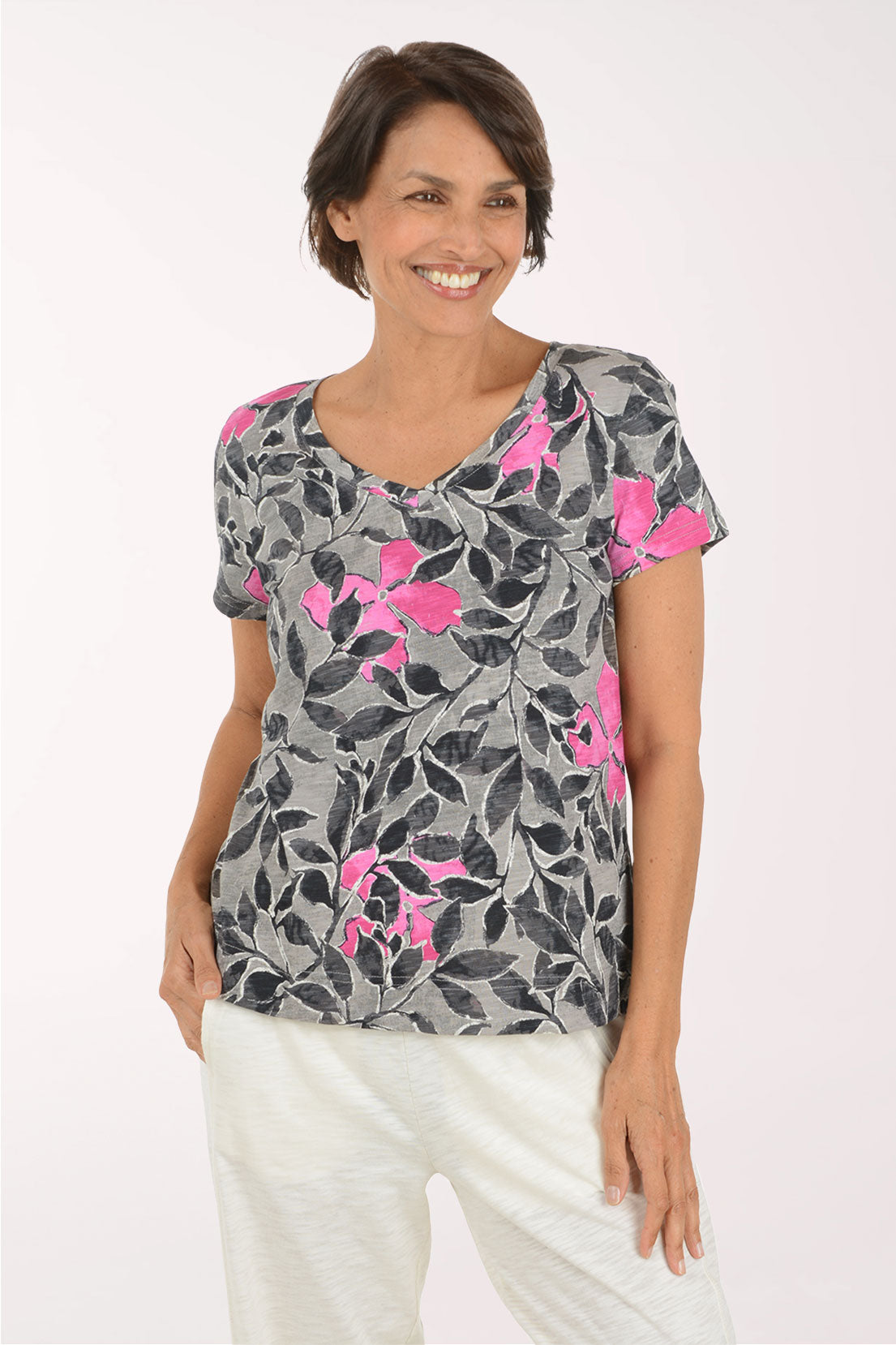 A woman wearing a gray short-sleeve V-neck top with a black leaf pattern and bright pink floral accents, paired with white pants, against a plain light background.