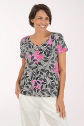 A woman wearing a gray short-sleeve V-neck top with a black leaf pattern and bright pink floral accents, paired with white pants, against a plain light background.