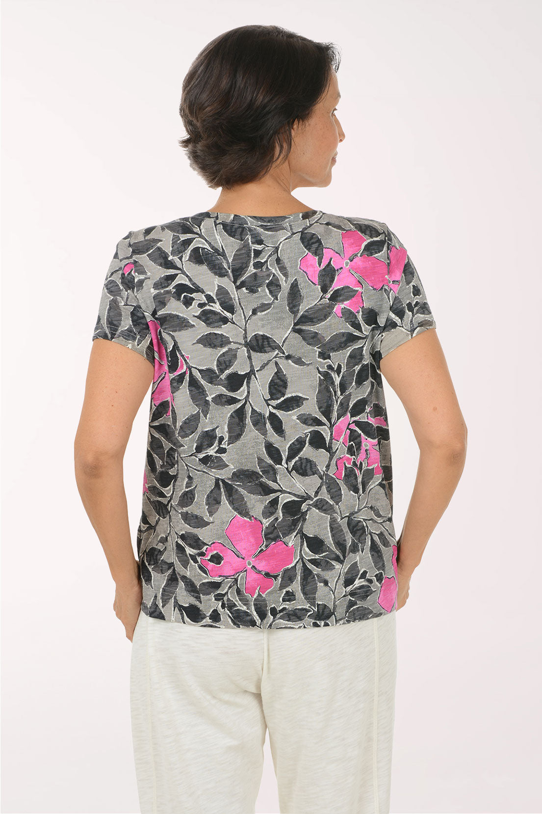 A woman wearing a gray short-sleeve V-neck top with a black leaf pattern and bright pink floral accents, paired with white pants, against a plain light background.